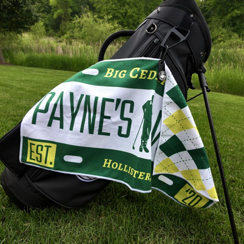 A custom golf towel featuring the text "PAYNE'S," "Big Cedar," and "Hollister" is clipped to a black golf bag standing on a green golf course. The towel includes a golfer silhouette and green, white, and yellow argyle accents. Trees and tall grass line the background.