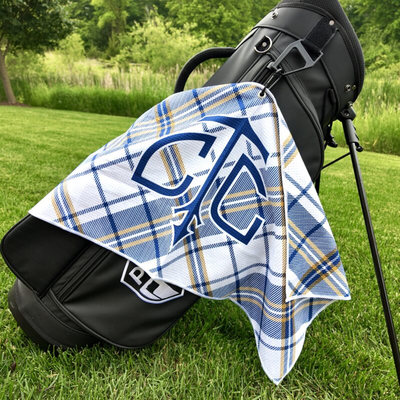 A black golf bag stands on a grassy area with a white towel draped over it. The towel features a navy, gold, and white plaid pattern with a large navy interlocking "C" and arrow logo at the center. Trees and tall grass fill the background.