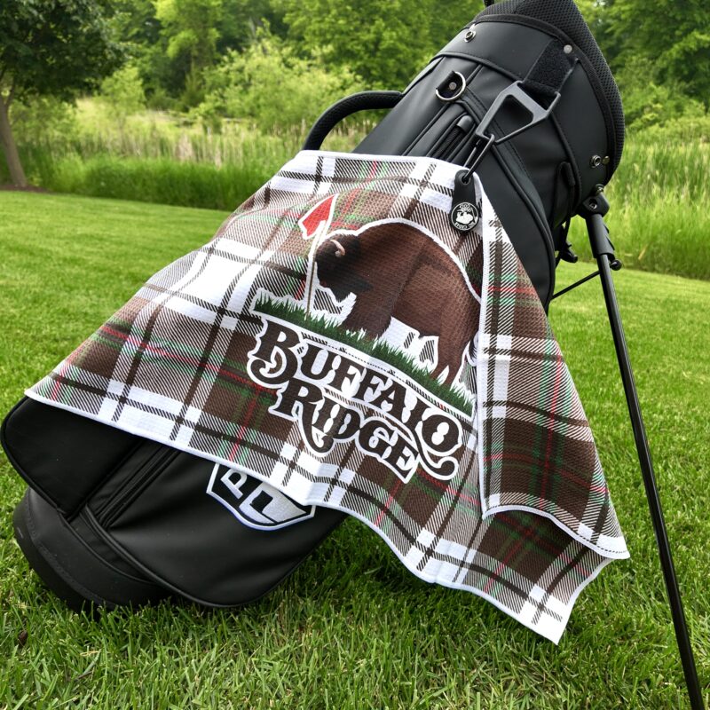 A black golf bag stands on vibrant green grass with a brown plaid towel hanging from it. The towel features a large graphic of a buffalo next to a red golf flag and the words “Buffalo Ridge” in bold white lettering. Tall grass and trees fill the background.