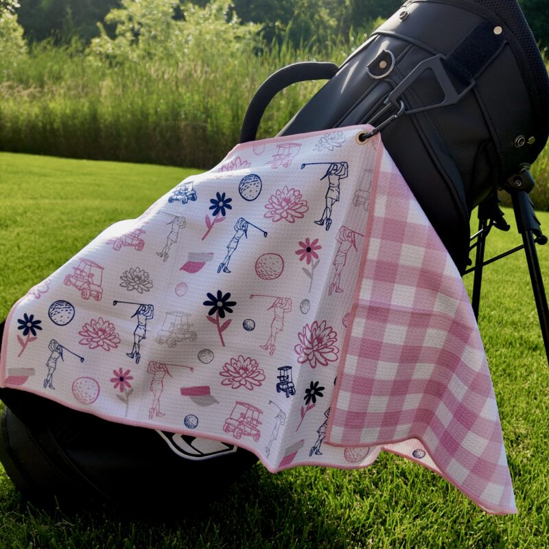 A black golf bag sits on vibrant green grass with a pink and white towel hanging from it. The towel features playful golf-themed illustrations, including golf carts, flowers, golf balls, and female golfers in pink, navy, and white, with a pink gingham pattern on one side. Tall grass and trees are visible in the background.