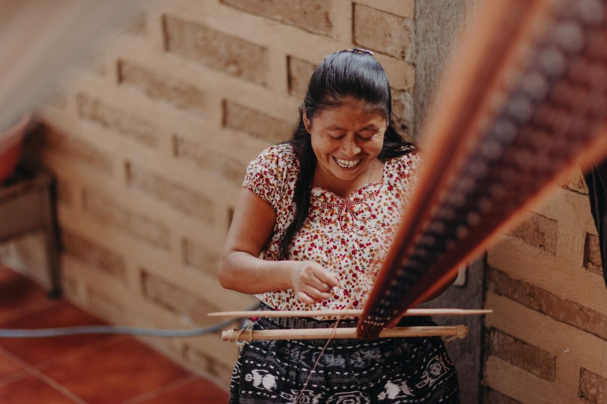 Smiling woman seated indoors using a traditional backstrap loom to weave fabric. She wears a floral blouse and patterned skirt, with brick walls and tiled floor visible in the background.