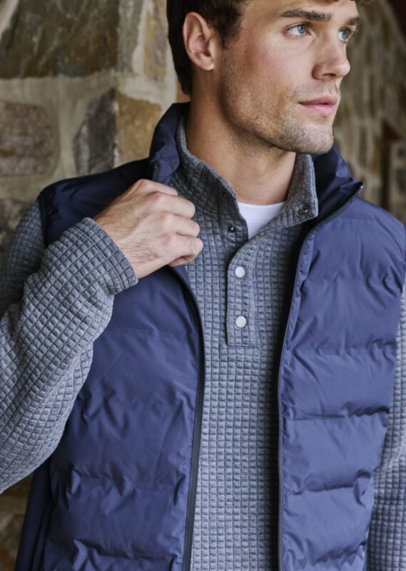 Man wearing a textured gray henley pullover layered with a navy puffer vest, standing in front of a stone wall and looking off to the side.