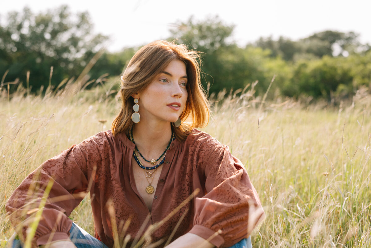 Woman sitting in a sunlit field of tall grass, wearing a rust-colored embroidered blouse, layered gold and blue necklaces, and large dangling earrings. She gazes thoughtfully into the distance, with her hair gently tousled by the breeze.