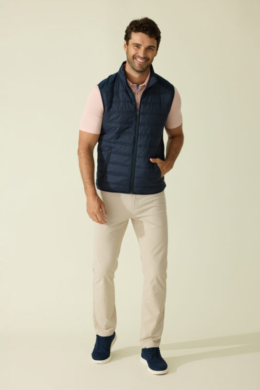 Man smiling and standing with one hand in his pocket, wearing a navy quilted vest over a light pink striped polo, beige pants, and navy sneakers against a light background.