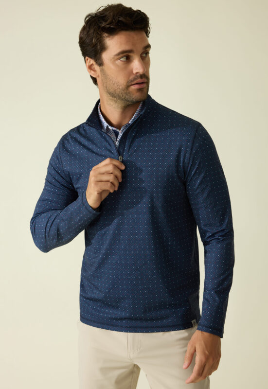 Man wearing a navy quarter-zip pullover with a subtle microdot pattern, layered over a collared shirt with a printed design, paired with light beige pants; he looks off to the side while adjusting the zipper.