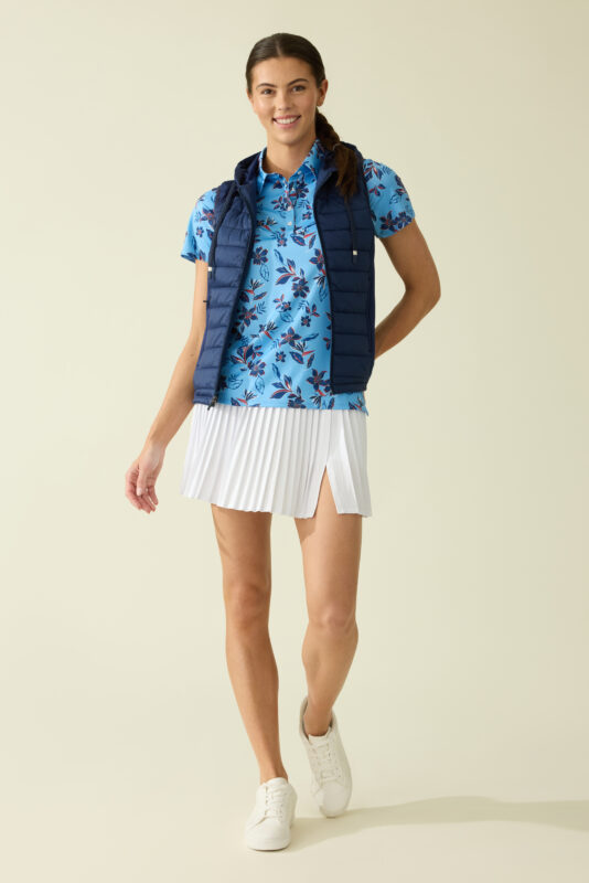 Woman modeling a blue floral short-sleeve golf polo layered under a navy quilted vest, paired with a white pleated skirt and white sneakers, standing against a neutral studio background.