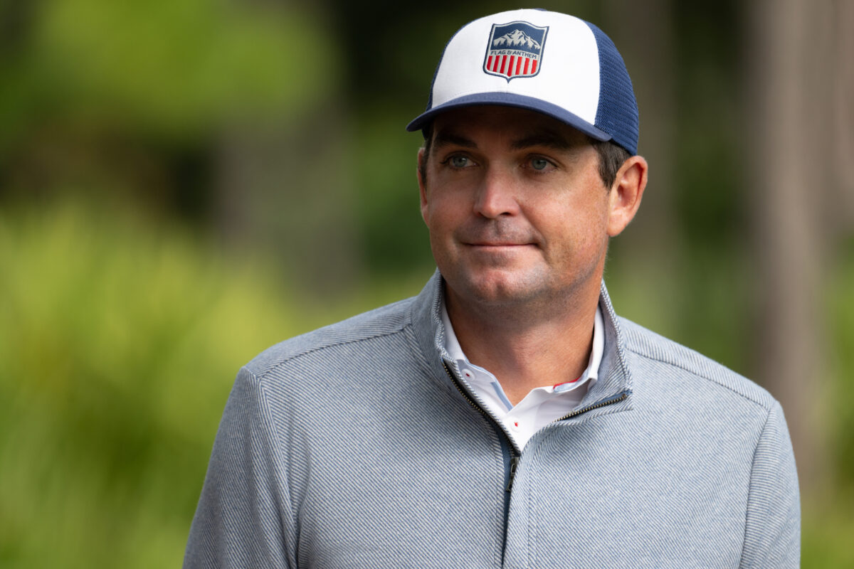 Man outdoors wearing a white and navy trucker hat with a red, white, and blue patch logo, and a light gray zip-up jacket over a collared shirt with a subtle red and blue print.
