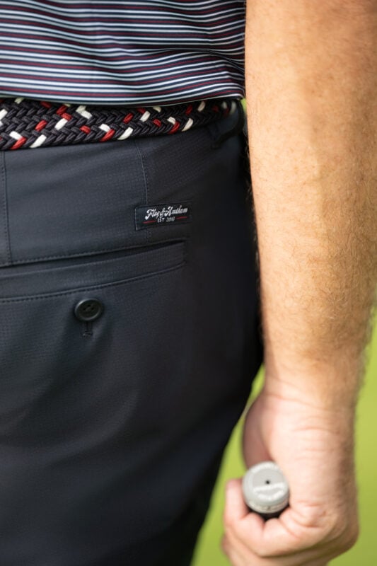 Close-up of a golfer’s navy performance pants with a back pocket and branded label, paired with a red, white, and navy braided belt and striped polo, holding a putter grip at his side.
