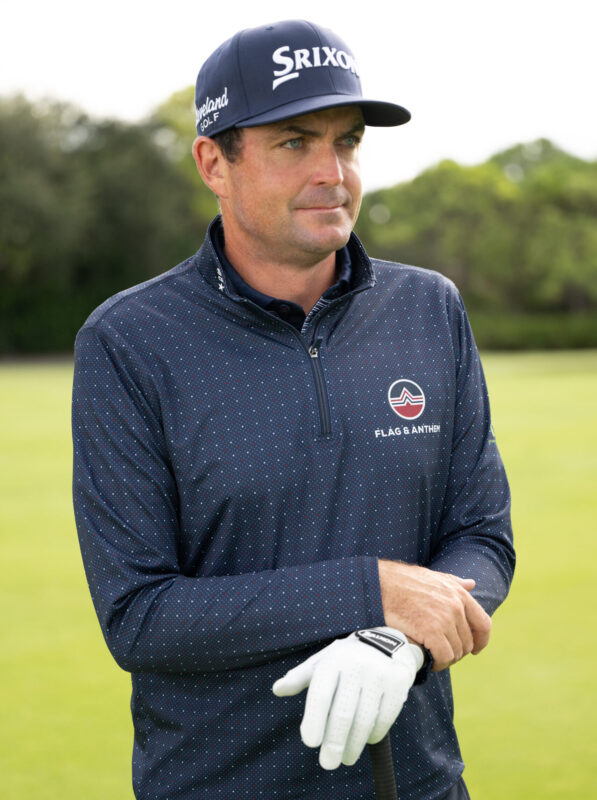 Male golfer wearing a navy Srixon hat and dotted quarter-zip pullover with "Flag & Anthem" logo, holding a golf club while standing on a green fairway.