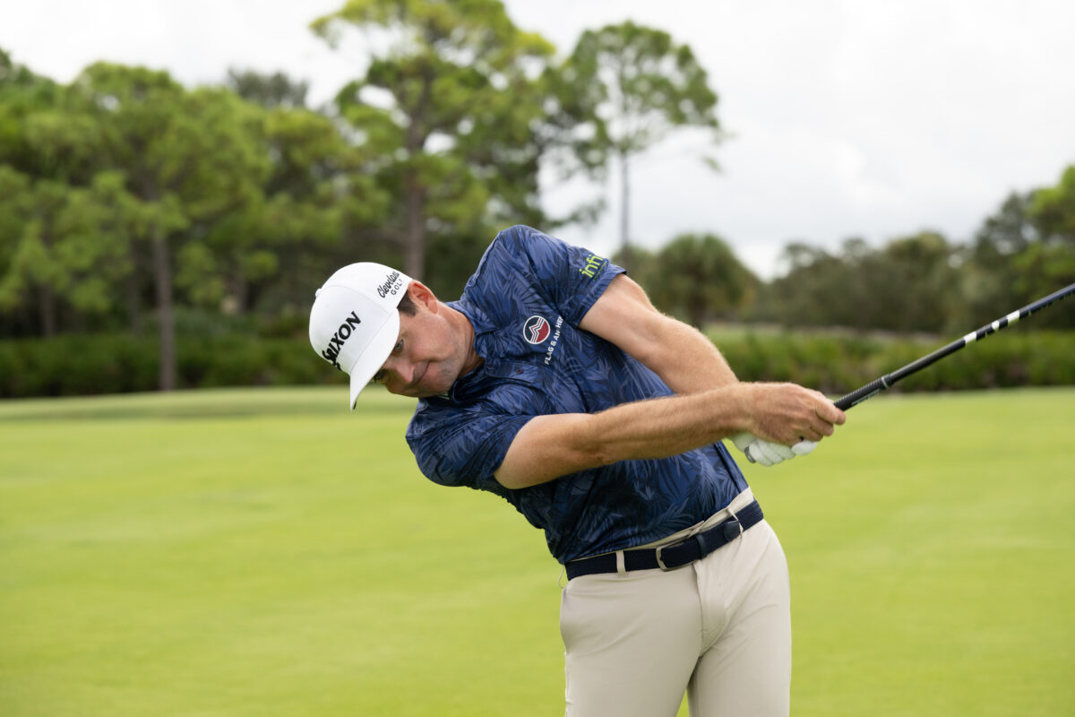 Male golfer in a navy patterned polo and light khaki pants completing his swing on a lush green course, wearing a white cap and white glove with trees in the background.