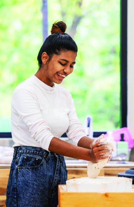 Young woman smiling as she works with clay in a brightly lit studio, wearing a white long-sleeve shirt and high-waisted jeans, with greenery visible through the window behind her.