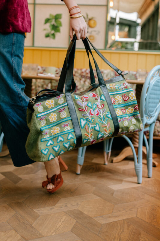 Close-up of a woman walking indoors holding a large olive green duffel bag with colorful embroidered patterns of hearts, flowers, and leaves. The bag features black leather straps, and the woman is wearing wide-leg jeans, rust-colored platform heels with bows, and gold bangles.