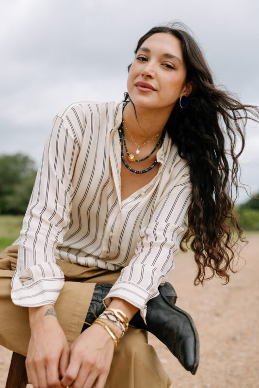 A woman with long, wavy dark hair poses outdoors on a wooden stool, wearing layered gold and blue necklaces, bold blue earrings, stacked bracelets, a striped button-up shirt, and tan wide-leg pants. One leg rests casually on the stool, revealing a black cowboy boot, with a cloudy sky and trees in the background.