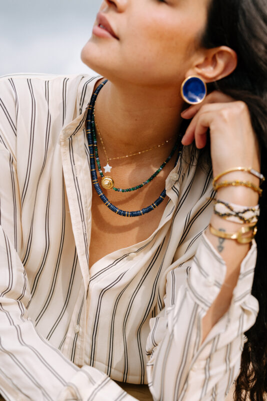 Close-up of a woman wearing layered necklaces in shades of gold, blue, and green, including a star charm and a gold medallion. She also wears a cream and black striped blouse, a large circular indigo earring, and a mix of gold and beaded bracelets. Her head is slightly tilted back, capturing a relaxed and elegant vibe.