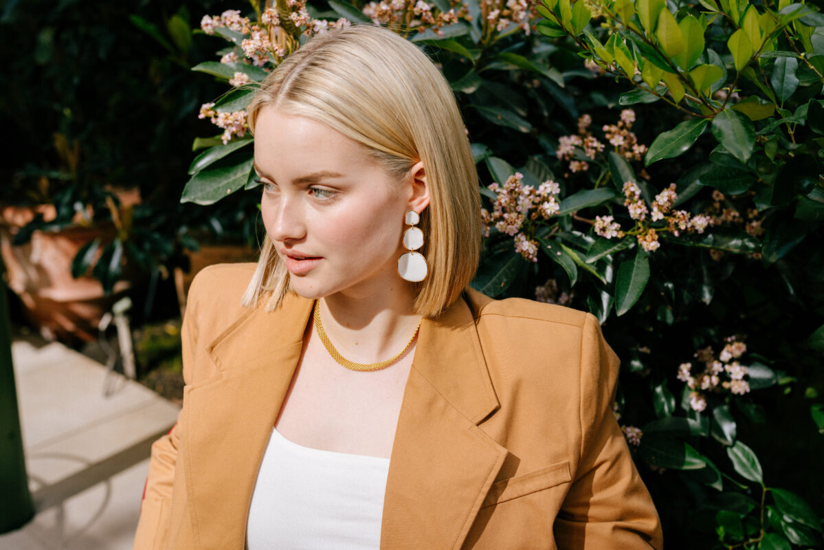 Blonde woman with a sleek bob hairstyle wearing bold white tiered statement earrings and a gold mesh necklace. She is dressed in a camel-colored blazer over a white top, seated in front of blooming greenery in bright natural light.