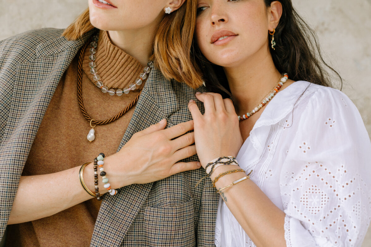 Two women standing close together, showcasing layered necklaces and bracelets. One wears a brown turtleneck and plaid blazer with a glass bead necklace, twisted cord with a pearl pendant, and earthy-toned bracelets. The other wears a white embroidered blouse, colorful beaded necklace, gold hoop earrings, and stacked woven and metal bracelets.