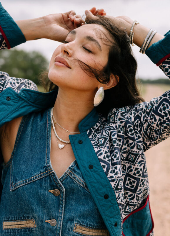 Woman outdoors with eyes closed and hands in her hair, wearing a patterned teal quilted jacket over a denim top, layered silver necklaces including a heart pendant, stacked bracelets, and large textured silver statement earrings.
