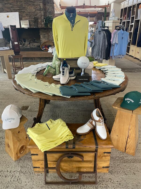 Golf shop display featuring a round wooden table with neatly arranged men's golf apparel in pastel greens and blues, centered by a mannequin dressed in a yellow quarter-zip and teal polo. Surrounding items include golf shoes, Callaway caps, a large Titleist golf ball prop, and folded shirts. Rustic wood elements and a stone fireplace create a cozy, upscale shop ambiance.