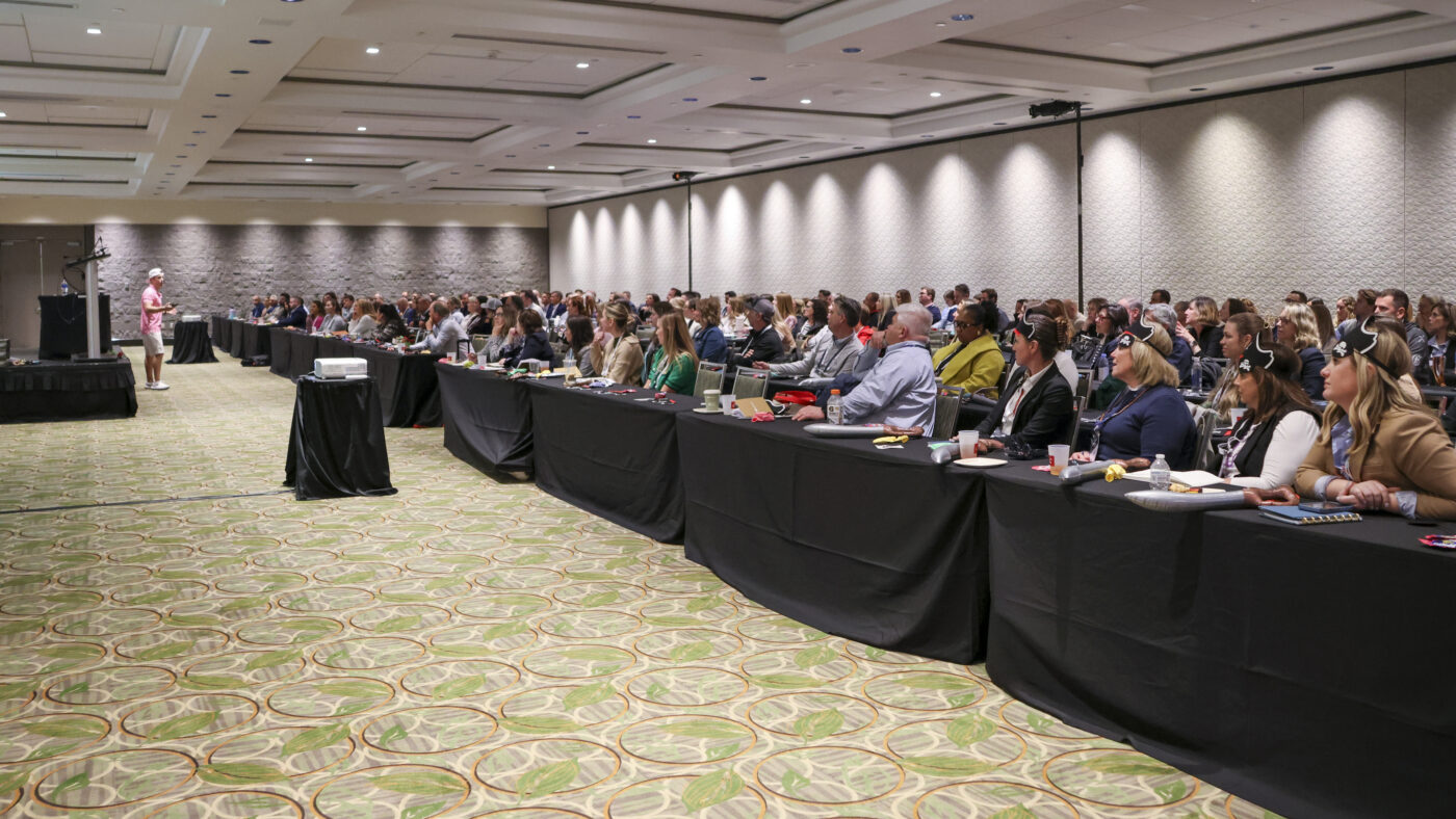 A large conference room filled with seated attendees listening attentively to a keynote speaker in a pink shirt and white cap at the front. Several attendees in the front row are wearing black pirate hats, adding a playful touch to the professional setting.
