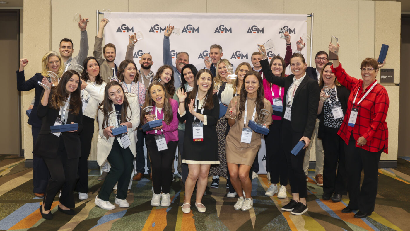 A large group of smiling AGM CRM graduates pose in front of an AGM-branded backdrop, proudly holding up their glass awards and certificate boxes. Many have lanyards and name tags, and the group appears energetic and celebratory.