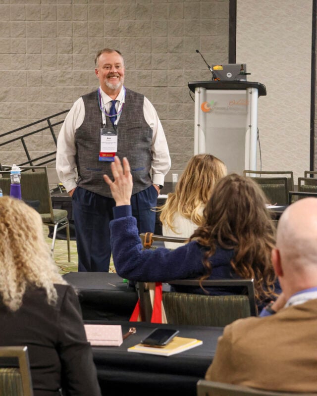 Ken, a smiling speaker wearing a vest and conference badge, stands at the front of a breakout room during a session. Audience members are seated at tables, one person in the foreground raising their hand to ask a question.