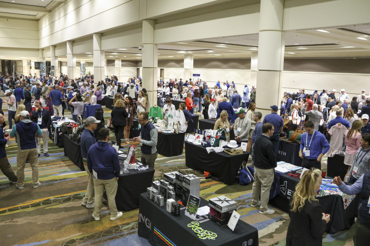 A large crowd of golf retailers, vendors, and buyers mingle at the AGM Product Preview Reception, walking through rows of display tables featuring apparel, accessories, and golf-related merchandise in a spacious, brightly lit convention hall.