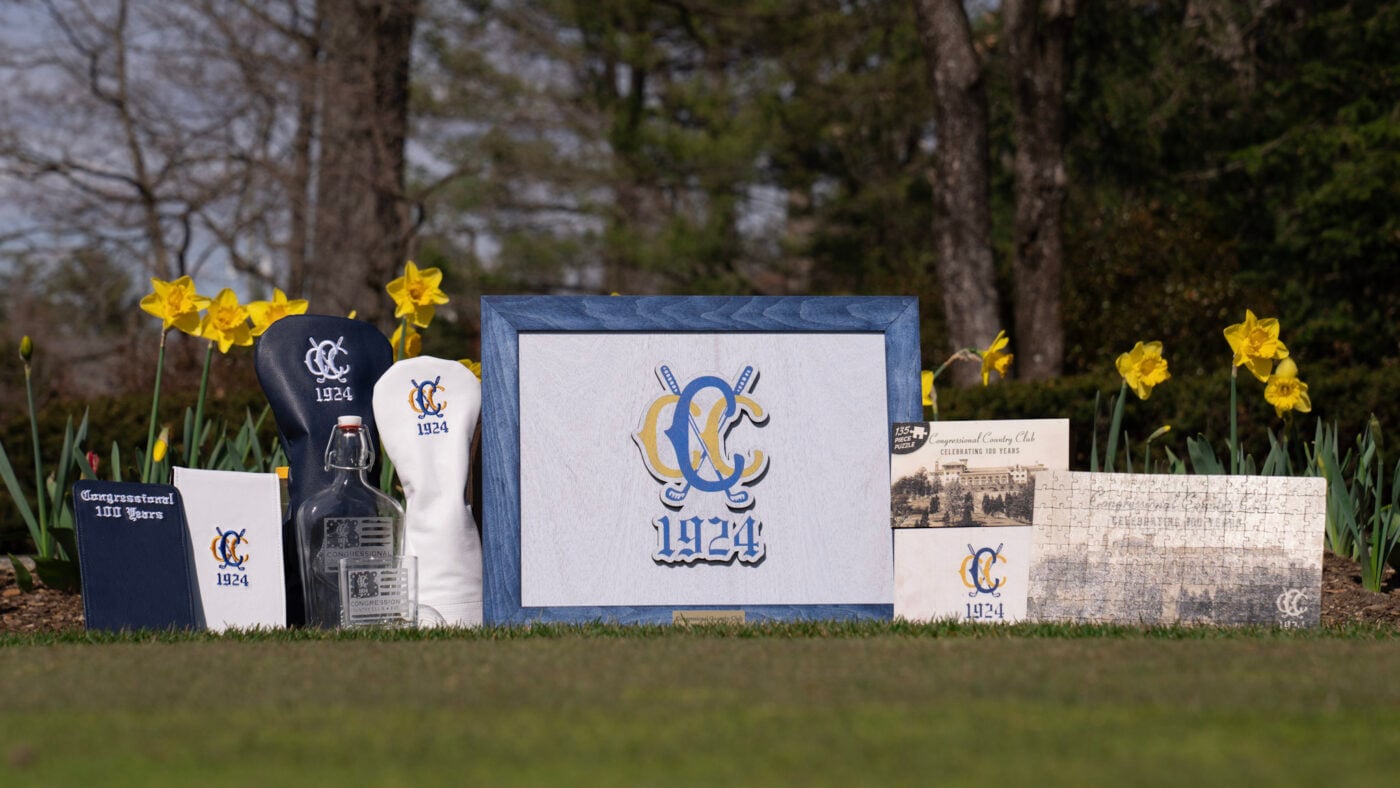 Outdoor display of Congressional Country Club centennial merchandise, including branded headcovers, a framed 1924 logo sign, a custom puzzle, leather accessories, a clear glass bottle, and embroidered towels. The items are arranged on grass in front of blooming yellow daffodils, with trees in the background.