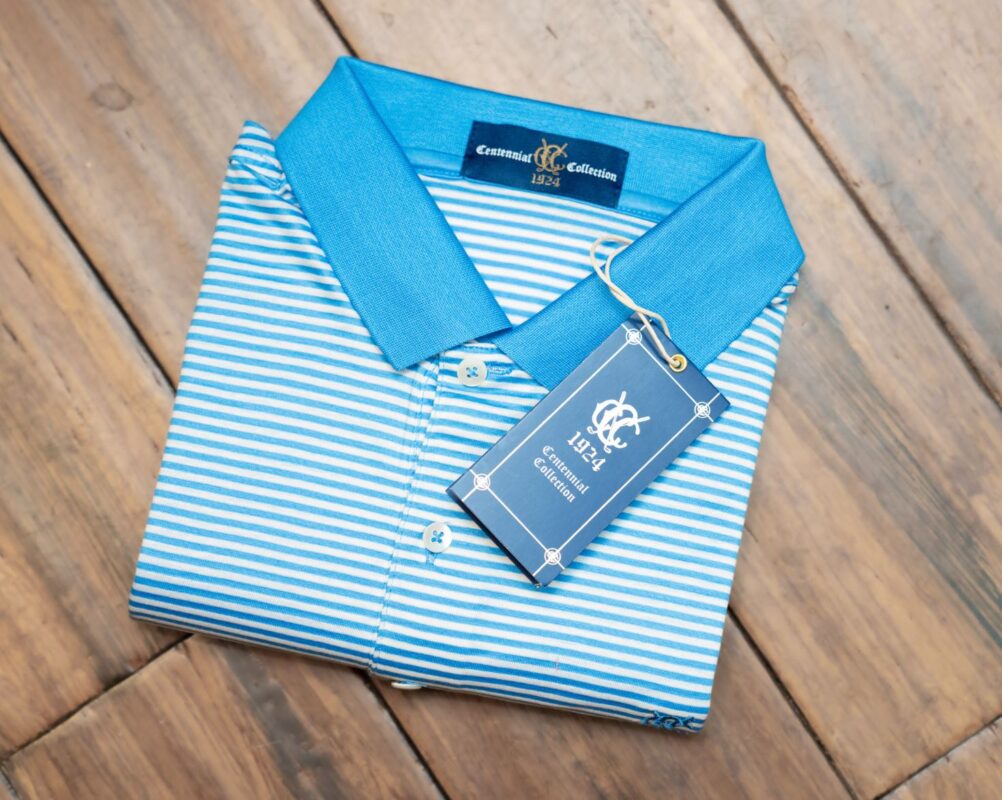 Folded light blue and white striped polo shirt with a solid blue collar, part of the Congressional Country Club 1924 Centennial Collection, displayed on a wooden floor with a navy blue hang tag featuring the club’s logo and branding.