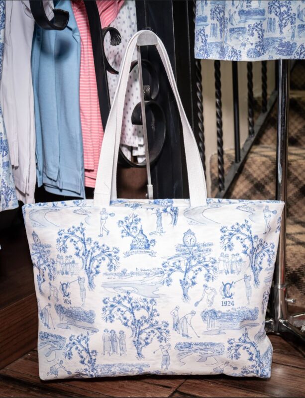 White canvas tote bag with blue toile-style illustrations of golfers, trees, and clubhouse scenes, featuring a club monogram and “1924,” displayed on a wooden floor beside a clothing rack in a retail setting.