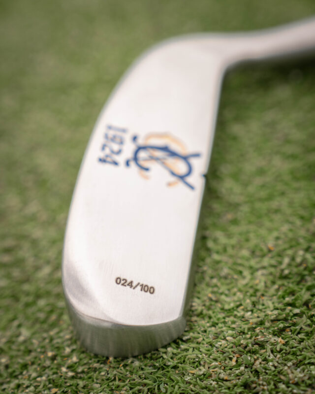 Close-up of a custom silver putter resting on green turf, engraved with a blue and gold club monogram and “1924,” with the limited-edition number “024/100” etched near the toe.