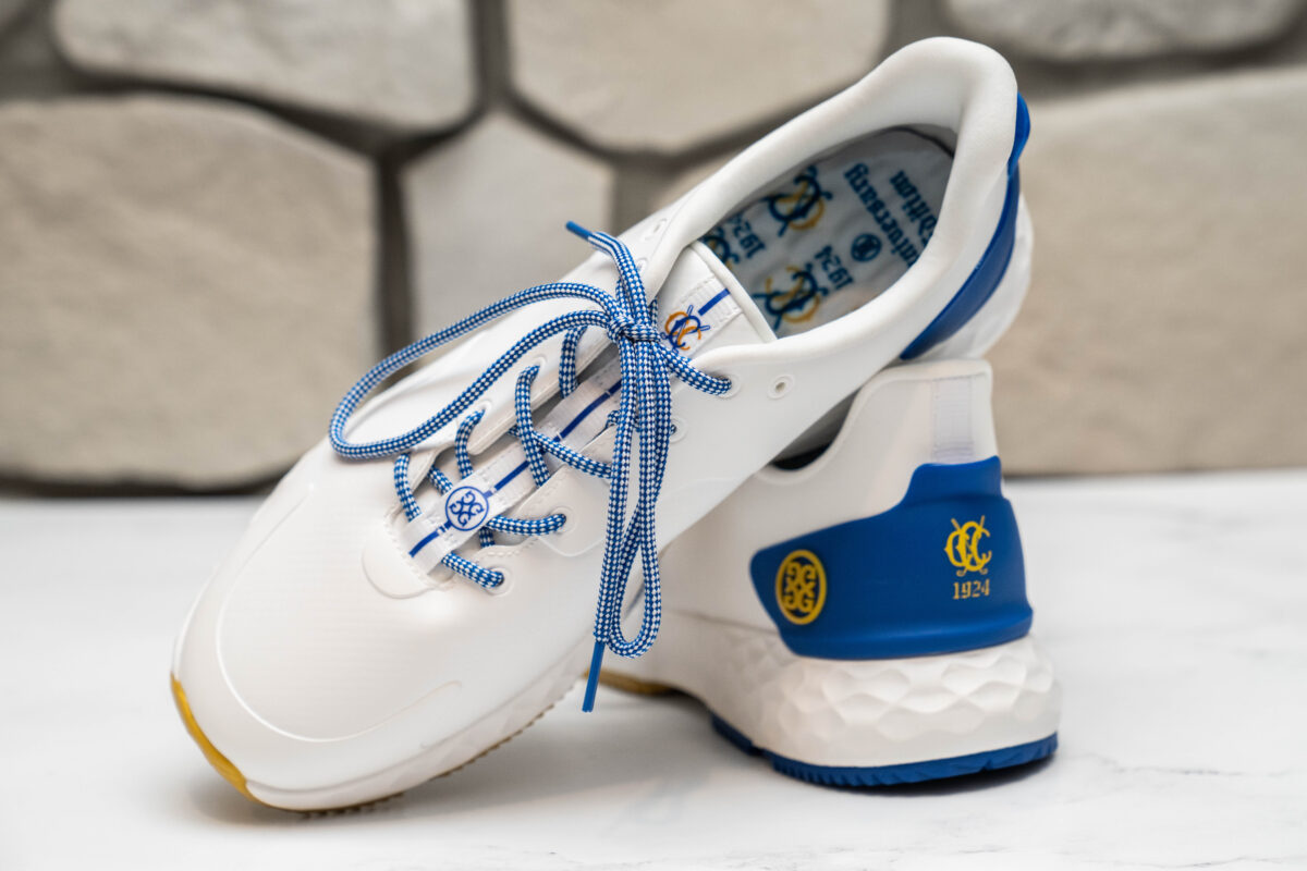 Close-up of a pair of custom white golf shoes with blue accents, blue-and-white checkered laces, and yellow details featuring the Congressional Country Club logo and "1924" on the heel. The shoes are displayed on a white surface with a stone wall background.