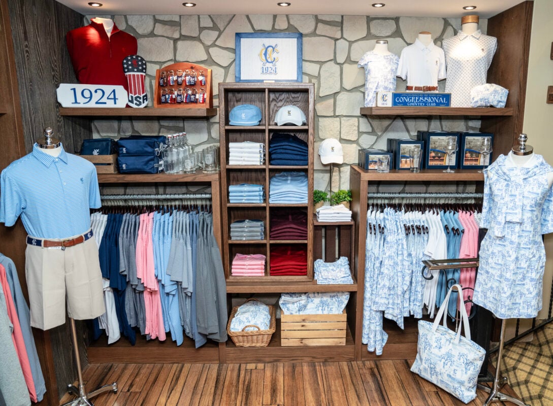 Merchandise display inside the Congressional Country Club golf shop featuring neatly folded polos, hats, and accessories in red, white, blue, and soft pastel colors. The display includes men's and women's apparel, branded items with “1924” and the club’s logo, and patriotic USA-themed headcovers, all arranged on wooden shelves against a stone wall backdrop.