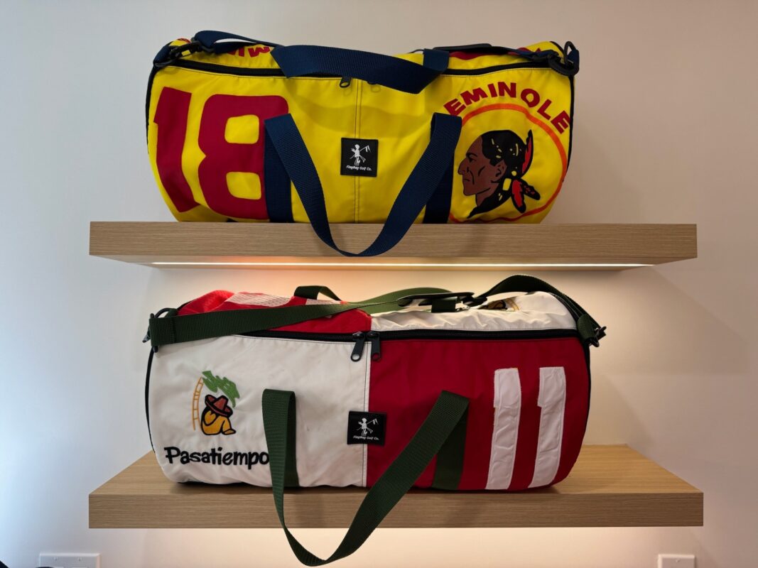 Two colorful duffel bags from FlagBag Golf Co. are displayed on wooden floating shelves. The top bag features a bold yellow and red design with "EMINOLE" and an illustrated Native American logo, while the bottom bag showcases a red, white, and green color scheme with "Pasatiempo" and a golfer under a tree. Both bags are made from repurposed golf course flags and include sturdy straps and zippers.