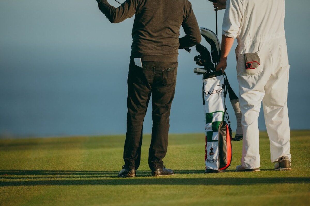 Two men stand on a golf course overlooking the ocean, one in a black jacket and pants, and the other in white caddie overalls. Between them is a custom white golf bag with green and red accents, embroidered with “Spyglass Hill” and “Pacific Grove Golf Links,” along with a lighthouse graphic.