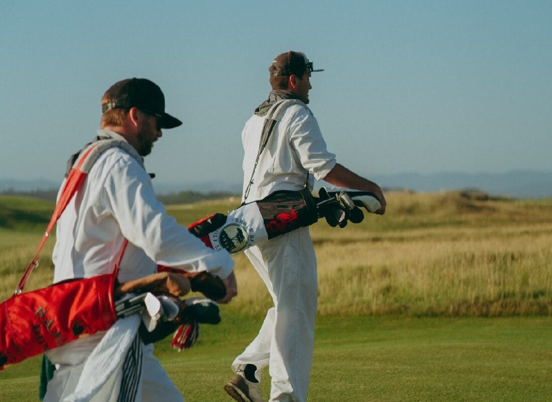 Two men dressed in white caddie uniforms walk across a sunny golf course carrying vintage-style FlagBag Golf Co. golf bags, with rolling hills and a clear blue sky in the background.