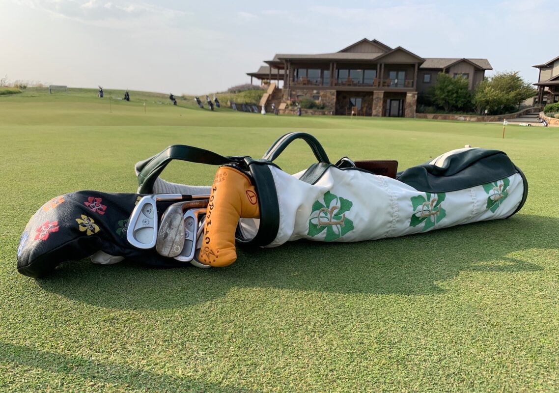 A custom white and green golf bag featuring a four-leaf clover design lies on the putting green with several clubs and colorful headcovers visible. In the background, a large clubhouse and several golfers can be seen practicing under a bright sky.