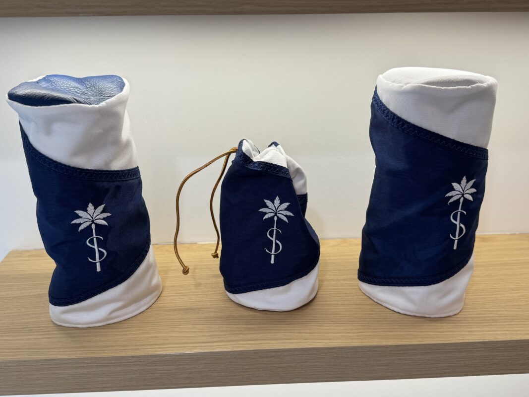 A set of three custom golf headcovers is displayed on a wooden shelf. Each headcover features a navy blue and white color scheme with a stitched palm tree and S emblem. The center cover is a smaller drawstring pouch, while the two on either side are standard-sized covers, likely for woods or drivers.