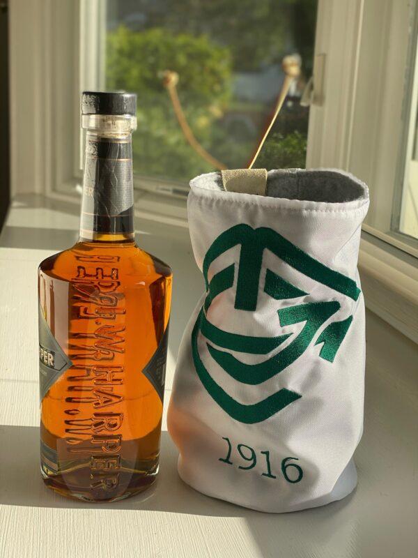 A bottle of I.W. Harper bourbon sits on a sunlit windowsill next to a white and green golf flag cozy embroidered with a stylized logo and the year “1916.” The window behind reveals a leafy outdoor scene blurred in the background.