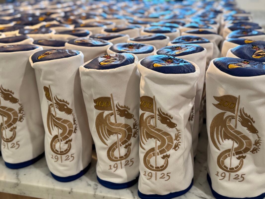 Rows of white golf headcovers lined up on a marble surface, each embroidered with a gold dragon wrapped around a flag marked “100” and the year “1925,” with a navy top panel featuring a colorful stitched emblem.