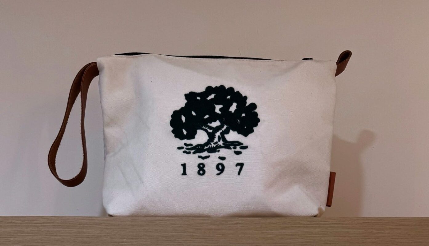 Close-up of a white pouch with a brown leather wrist strap, featuring a black embroidered oak tree and the year "1897" underneath, displayed on a wooden shelf against a neutral background.