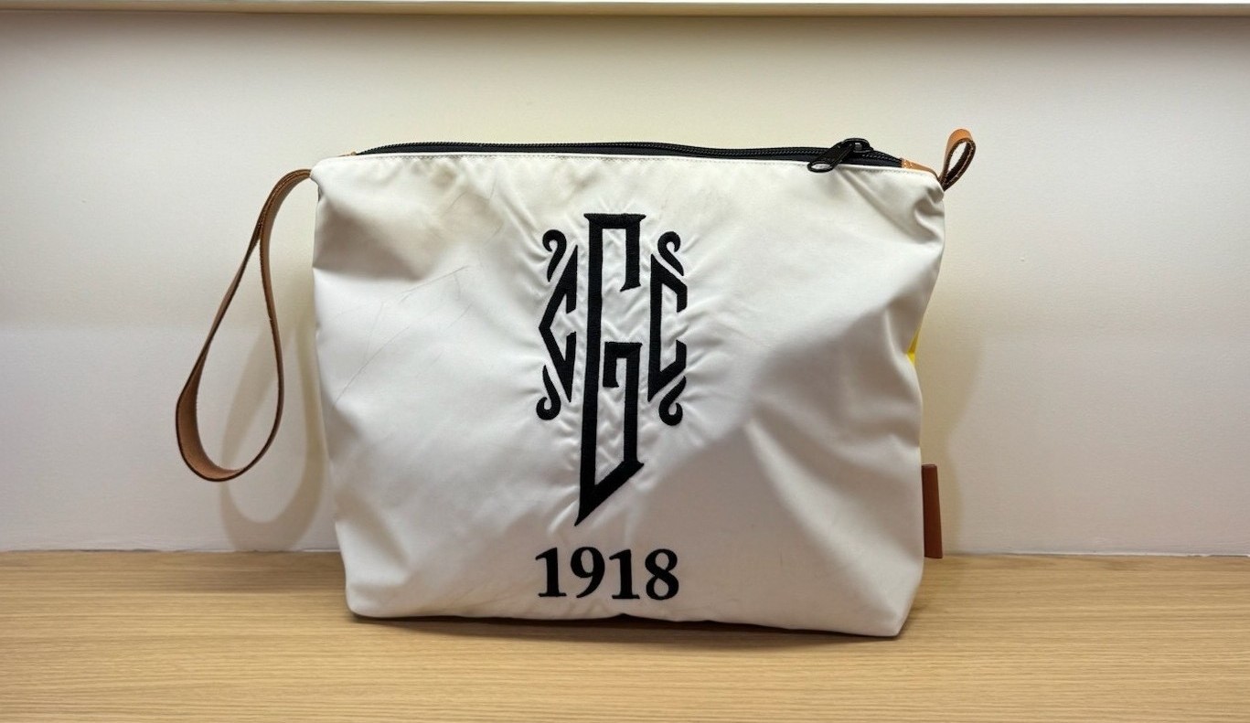 White zippered pouch with a black embroidered monogram-style logo and the year “1918” on the front, featuring a black zipper and brown wrist strap, displayed on a wooden shelf against a white wall.