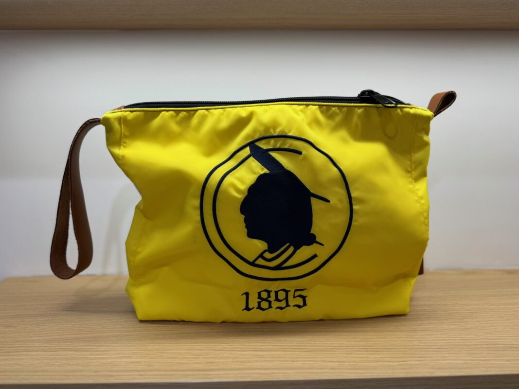 Bright yellow zippered pouch with a brown leather wrist strap, featuring a black embroidered silhouette of a Native American profile within a circular border and the year “1895” stitched below, displayed on a wooden shelf.