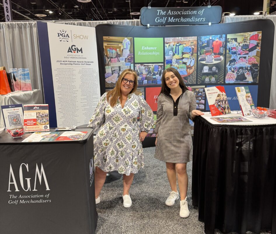 Two smiling women stand in front of the Association of Golf Merchandisers booth at the 2025 PGA Show. The booth features promotional materials, a Platinum Awards poster, and colorful retail display photos with messaging like “Enhance Relationships” and “Elevate Retail.” One woman wears a patterned dress and white slip-ons, the other wears a black-and-white checkered dress and white sneakers.