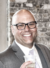 Steve Risler headshot. Bald man wearing black glasses smiling into the camera and carrying a drink. He is wearing a white collared shirt and grey blazer and has a stone background.