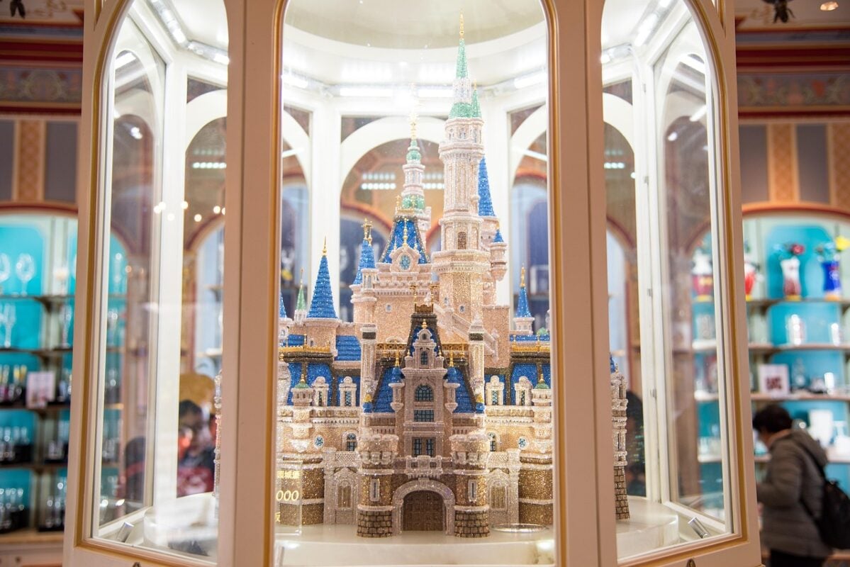 A sparkling miniature replica of a Disney castle, intricately detailed and encrusted with crystals, displayed inside a glass case with white framing. The background shows a store interior with shelves of glassware and colorful merchandise, and a person browsing on the right side.