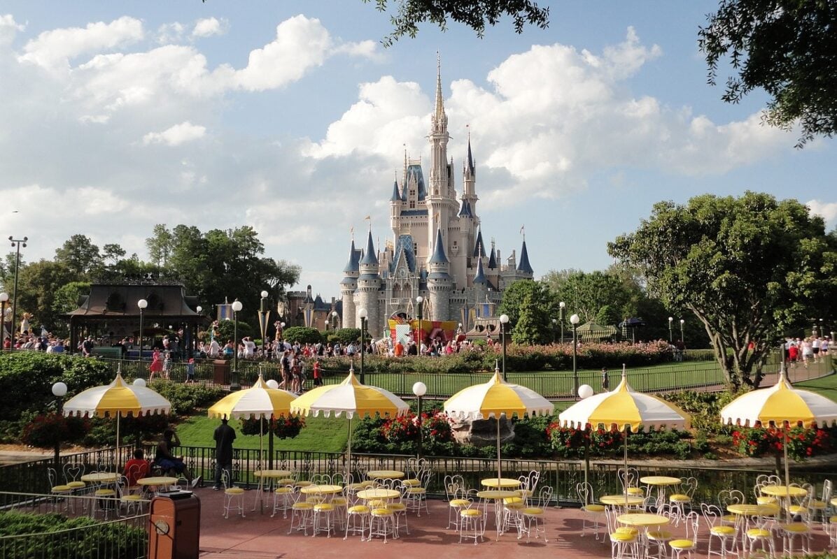 Wide view of Cinderella’s Castle at a Disney theme park, surrounded by lush gardens and crowds of visitors. In the foreground, yellow-and-white striped umbrellas shade an outdoor seating area with round yellow tables and white wrought iron chairs. The sky is bright with scattered clouds, adding to the cheerful atmosphere.
