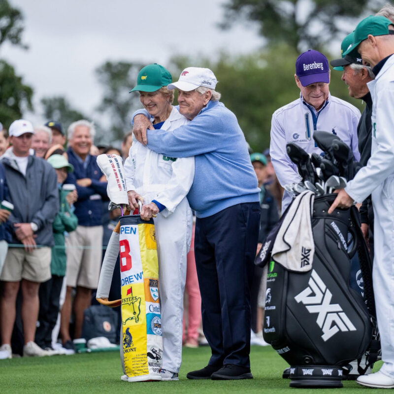 Jack Nicklaus embraces his wife, Barbara, on the tee box during the ceremonial start of a golf tournament. Barbara is dressed in the traditional white caddie uniform and holding a commemorative golf bag featuring patches from historic courses. Spectators and fellow golfers look on and smile, capturing a heartfelt and celebratory moment.