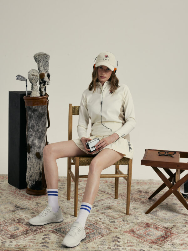 Female model wearing a cream long-sleeve golf top, matching skirt, and white cap, seated on a wooden chair while holding a portable music player and wearing headphones, with a vintage golf bag and clubs beside her in a studio setting.
