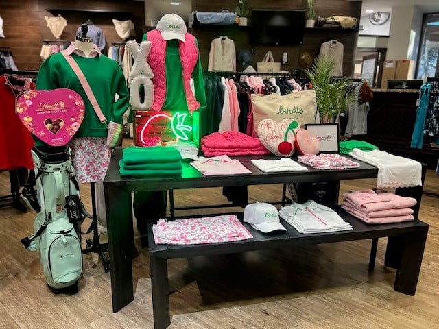 Valentine’s-themed golf shop display featuring mannequins in green and pink apparel, a mint golf bag, folded coordinated outfits on a black table, heart-shaped gift items, and festive décor including a neon heart accent and “XO” balloons.