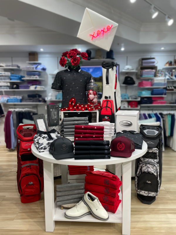 A circular white table at The Sanctuary Golf Club features a Valentine’s Day retail arrangement in red, black, and white. A mannequin bust in a black patterned polo is topped with red roses, surrounded by a red marquee "love" sign, heart ornaments, and a white mailbox. The display includes stacked performance polos, G/FORE hats, white golf shoes, and club-branded golf bags in red and camo. Hanging above the table is a white envelope prop with a glowing red neon "xoxo" sign.